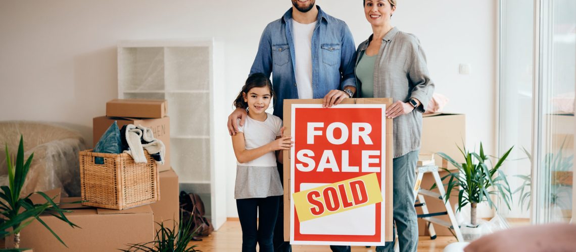 Happy family standing in their new home while holding sold real estate sing and looking at camera.